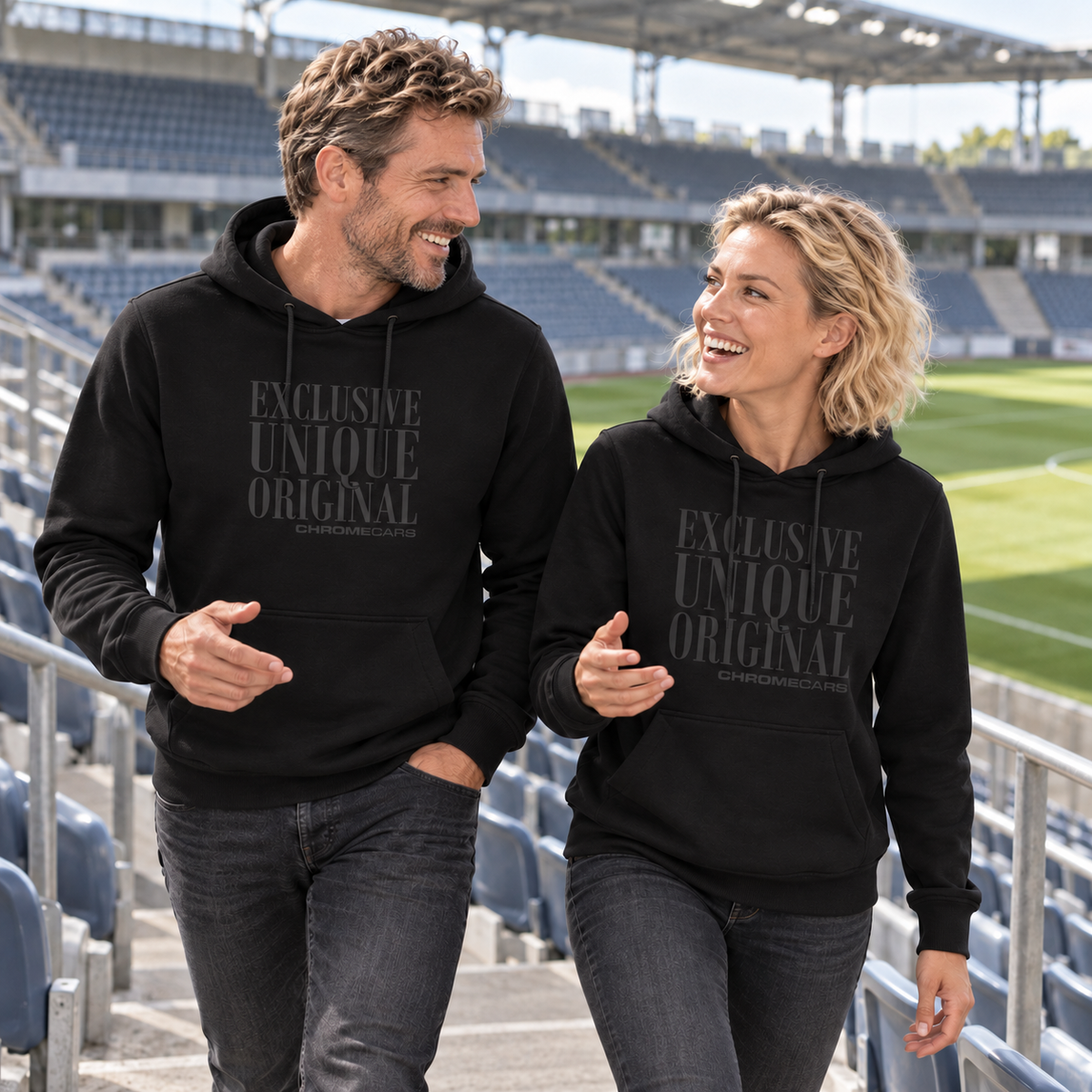 Ein Mann und eine Frau gehen in schwarzen Hoodies mit schwarzen Typografiemotiven nebeneinander durch einen Tribünengang im Fußballstadion, sehen einander an und gestikulieren mit jeweils einer Hand.
