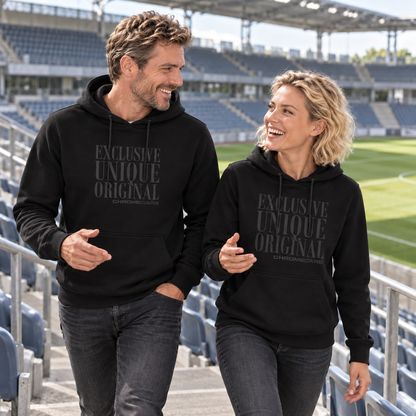 Ein Mann und eine Frau gehen in schwarzen Hoodies mit schwarzen Typografiemotiven nebeneinander durch einen Tribünengang im Fußballstadion, sehen einander an und gestikulieren mit jeweils einer Hand.