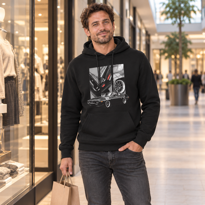 Mann im schwarzen Hoodie steht neben einem Schaufenster und hält eine Einkaufstasche. Vorn zeigt der Hoodie ein dunkles Coupé unter drei Detailfeldern.