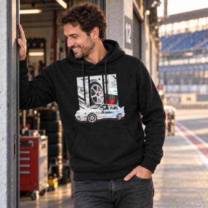 Mann im schwarzen Hoodie lehnt eine Hand an einen Garagentürrahmen in einer Boxengasse. Vorn zeigt der Hoodie ein weißes Auto mit türkisfarbener Grafik und drei Detailfelder.