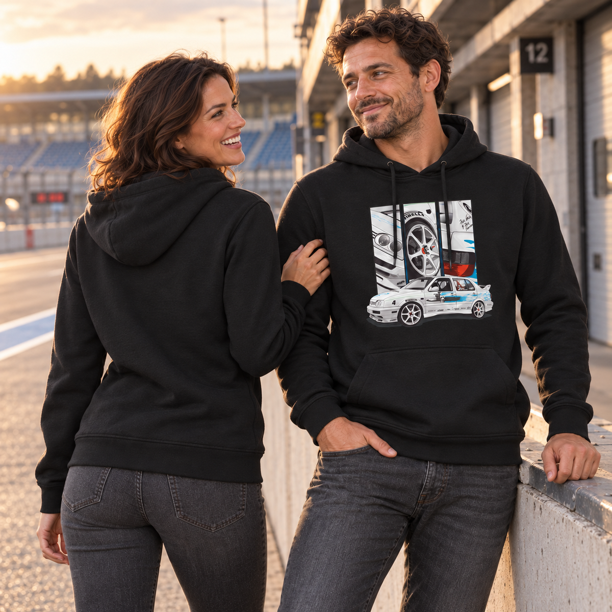 Paar in schwarzen Hoodies steht an einer Boxengasse. Die Frau zeigt den unbedruckten Rücken, der Mann das weiße Frontmotiv mit türkisfarbener Grafik.