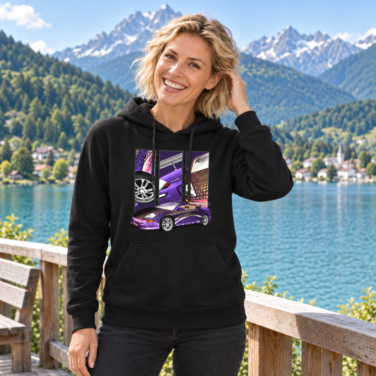 Frau im schwarzen Hoodie steht lächelnd am Bergsee und berührt ihr Haar. Ihre Vorderseite zeigt das violette Fahrzeugmotiv mit Gittergrafik und drei Feldern.