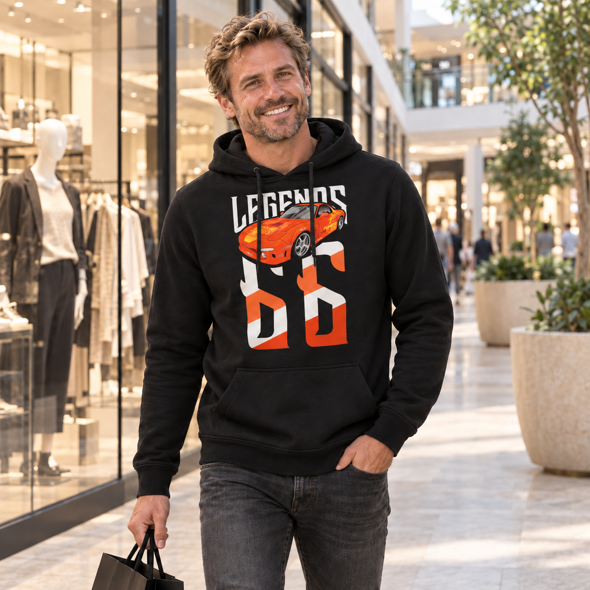 Mann im schwarzen Hoodie mit Orange Julius RX-7 als Legends-Frontmotiv geht mit einer Einkaufstasche durch ein helles Shopping-Center.