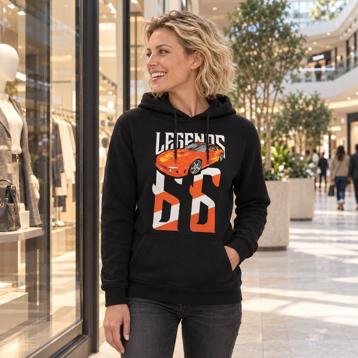 Frau im schwarzen Legends-Hoodie mit Orange Julius RX-7 auf der Vorderseite steht vor einem Schaufenster im hellen Shopping-Center.