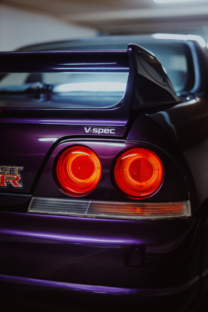 Vertikale Nahaufnahme des dunkelvioletten Hecks eines Nissan Skyline V-Spec mit zwei rot leuchtenden Rundrückleuchten, GT-R-Emblem, großem Heckflügel und blau-violetten Lichtreflexen vor unscharfem dunklem Hintergrund.