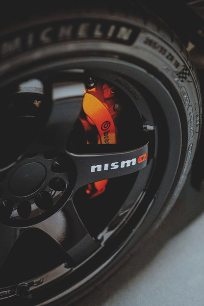 Nahaufnahme eines schwarzen Fahrzeugrads mit dunkler Mehrspeichenfelge, hellem Nismo-Schriftzug und orange-rotem Brembo-Bremssattel. Reifenflanke und Radmuttern rahmen das Motiv; körniges Licht, starke Schatten und geringe Schärfentiefe prägen den engen Hochformat-Ausschnitt.