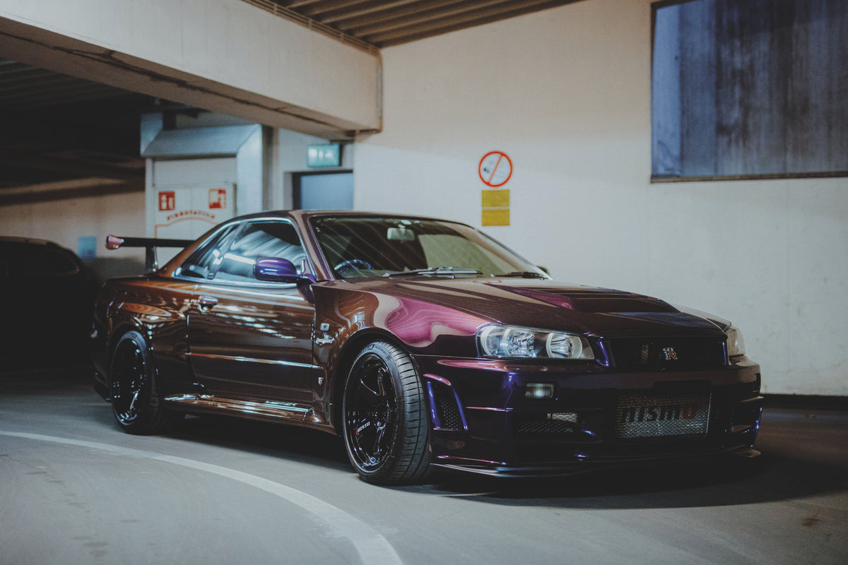 Violetter Nissan Skyline R34 schräg von vorn vor einer hellen Garagenwand und einer dunklen Öffnung.