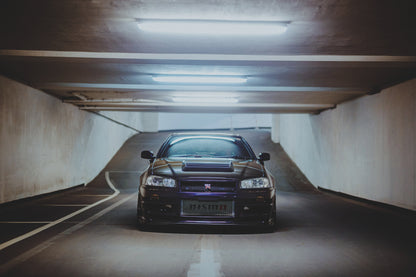 Dunkelvioletter Nissan Skyline R34 in symmetrischer Frontansicht auf einer Parkrampe. Helle Betonwände, Fahrbahnmarkierungen und kühle rechteckige Deckenleuchten rahmen das zentral stehende Fahrzeug; die Rampe steigt im Hintergrund an.