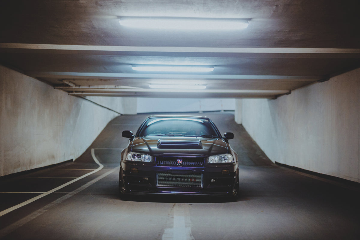 Dunkler Nissan Skyline R34 frontal über einer weißen Bodenlinie in einer beleuchteten Tiefgarage.