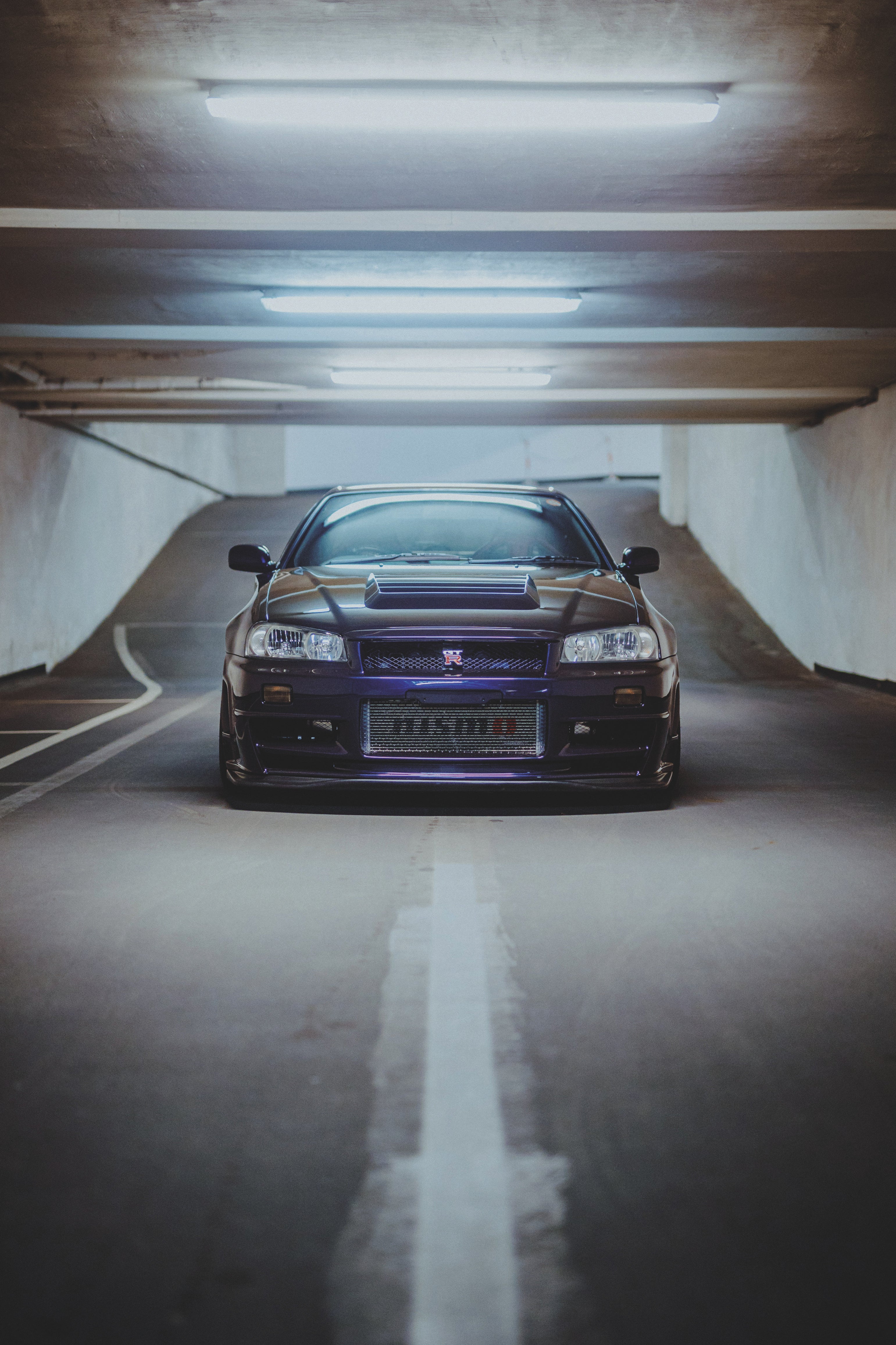 Dunkelvioletter Nissan Skyline R34 frontal in einer Tiefgarage, mittig über einer weißen Bodenlinie unter Deckenleuchten.