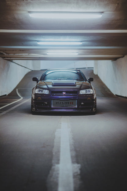 Dunkelvioletter Nissan Skyline R34 in zentraler Frontansicht auf der Zufahrt einer Parkgarage, beleuchtet von mehreren weißen Deckenleuchten und umgeben von grauen Wänden und Fahrbahnmarkierungen.
