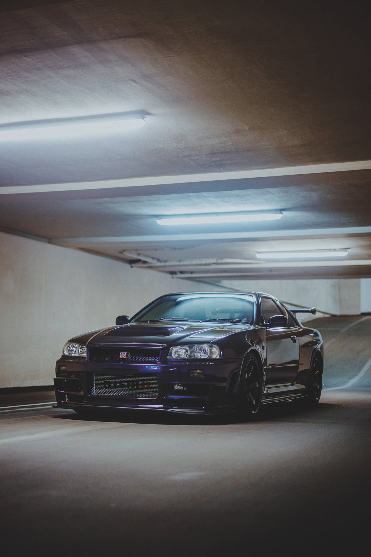 Dunkler Nissan Skyline R34 schräg von vorn in einer Tiefgarage unter langen Deckenleuchten.