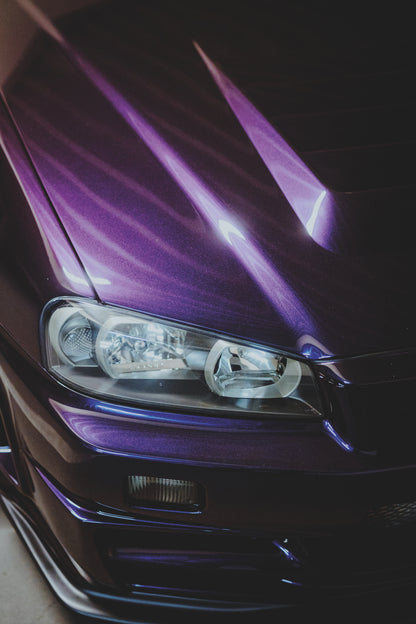 Nahaufnahme der dunkelvioletten Front eines Nissan Skyline R34, schräg von vorn fotografiert. Ein Scheinwerfer und Teile des Stoßfängers sind sichtbar; helle violette Reflexe verlaufen über die Motorhaube vor dunklem Hintergrund.