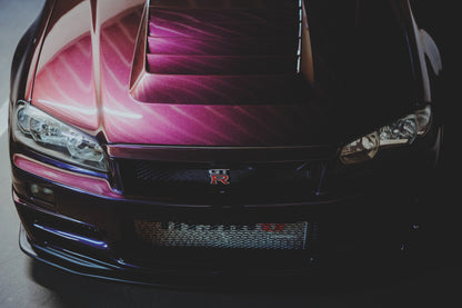 Enge Frontansicht eines dunkelvioletten Nissan Skyline R34 mit sichtbarem GT-R-Emblem, Haubenöffnungen und Scheinwerfern; magentafarbenes Licht links, tiefer Schatten rechts.