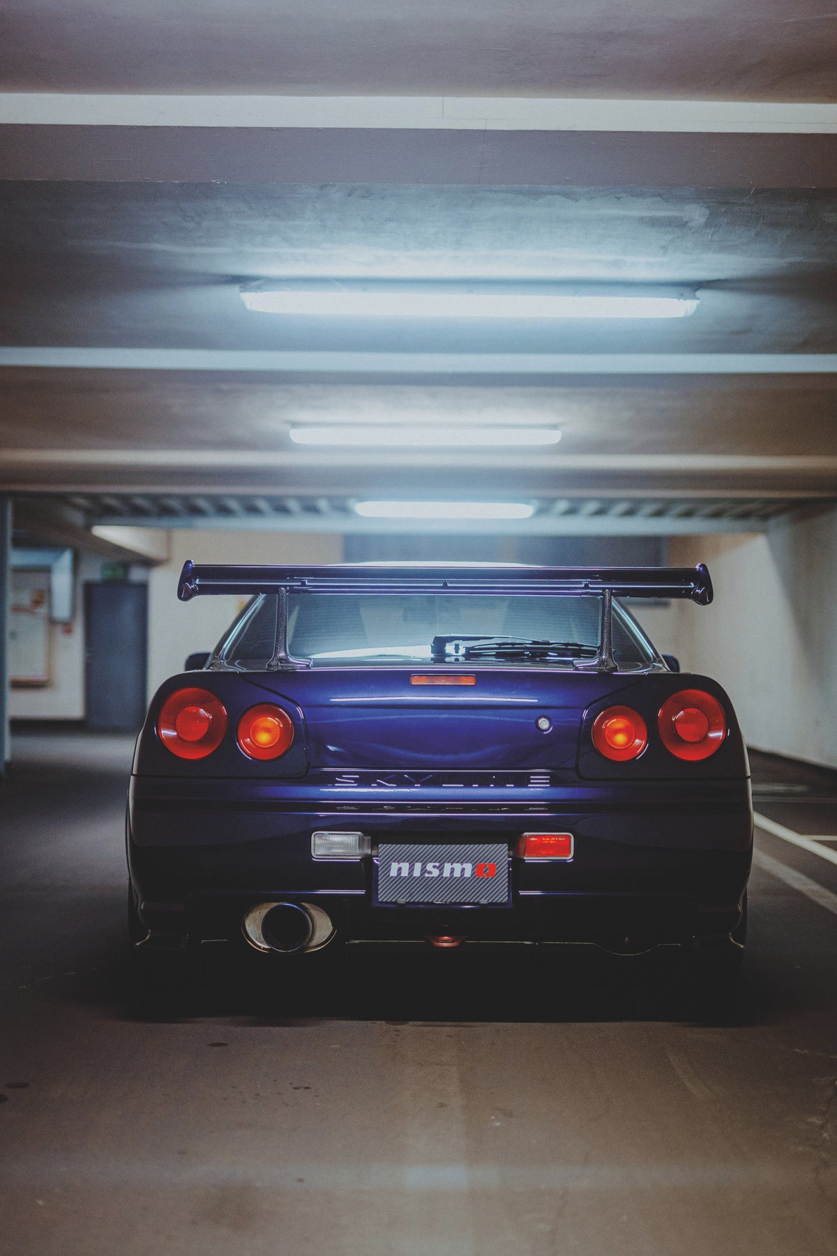 Gerade Heckansicht eines violetten Nissan Skyline R34 mit vier runden Rückleuchten unter Deckenleuchten.