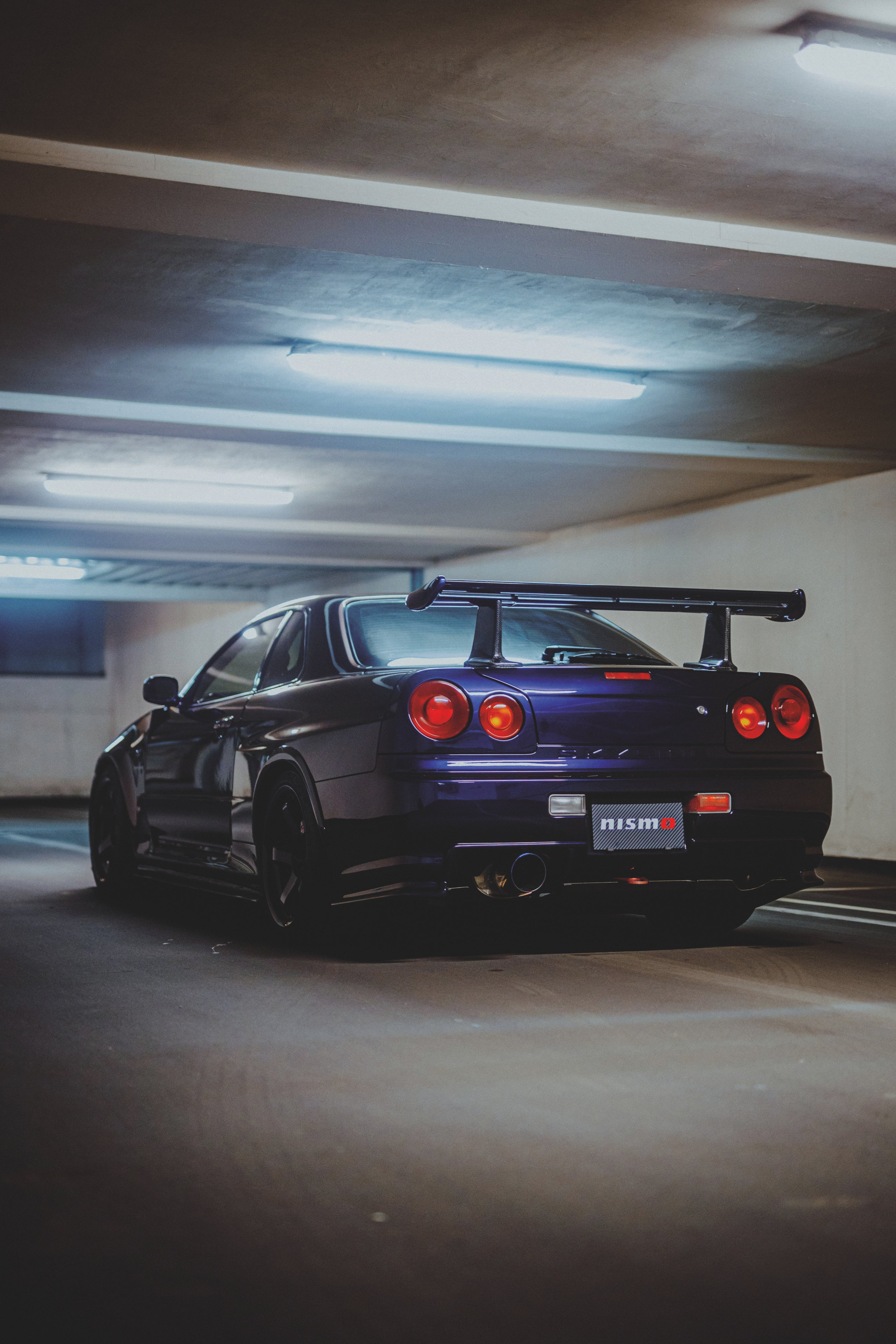 Violetter Nissan Skyline R34 schräg von hinten in einer Tiefgarage, mit vier runden roten Rückleuchten und großem Heckflügel.
