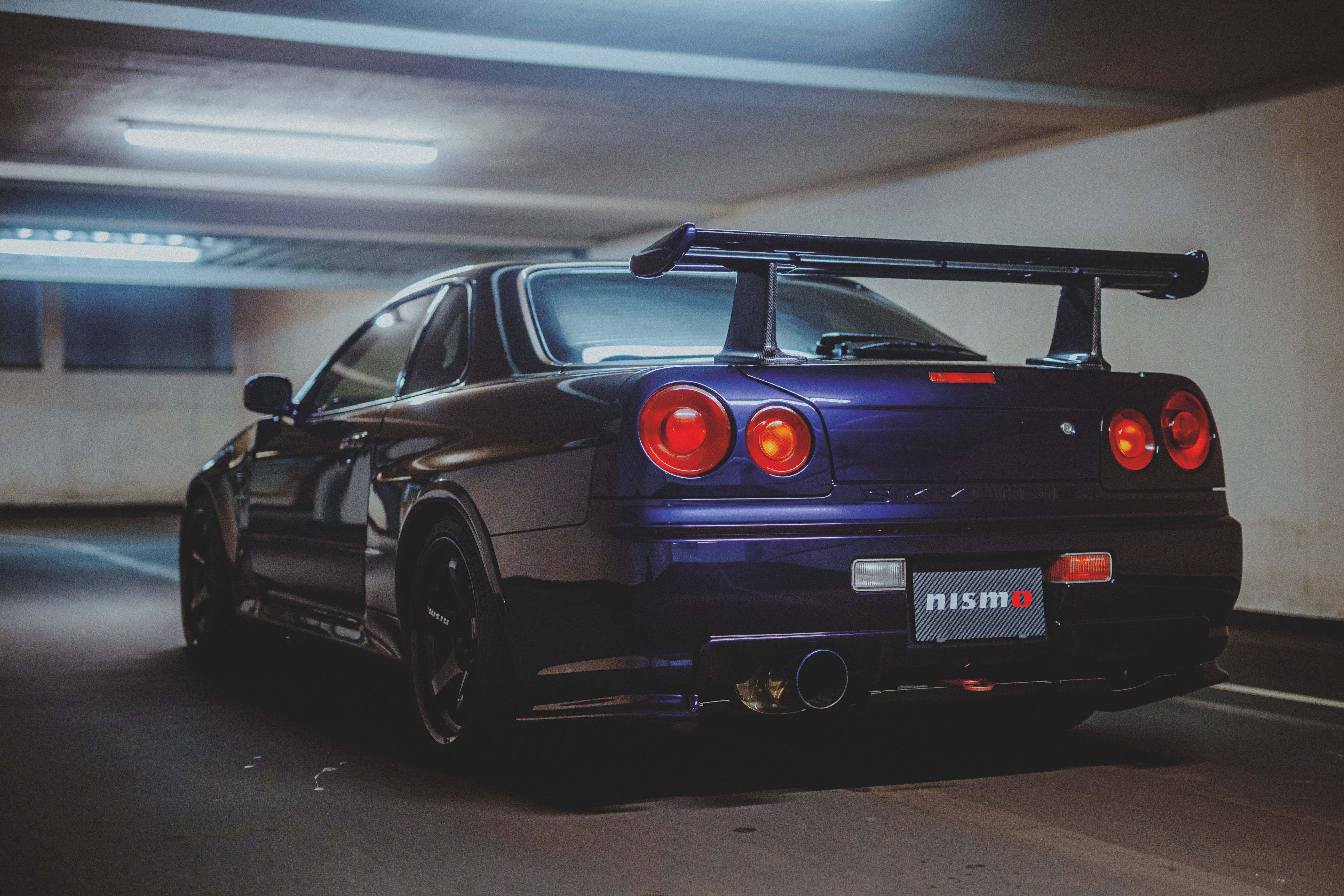 Violetter Nissan Skyline R34 schräg von hinten mit großem Heckflügel und vier runden Rückleuchten in einer Tiefgarage.