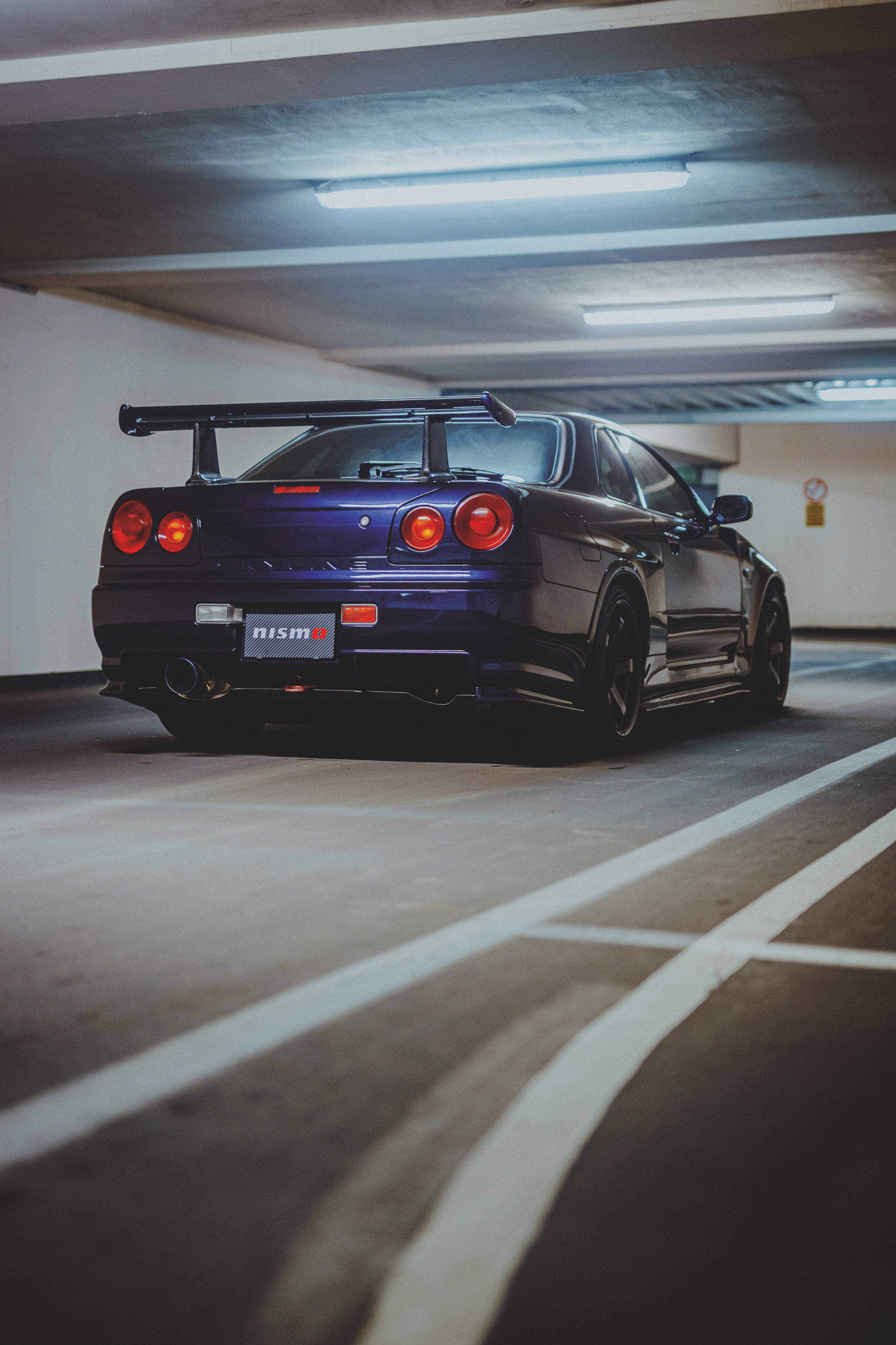Nissan Skyline R34 schräg von hinten neben weißen Bodenmarkierungen, mit Heckflügel und runden Rückleuchten.