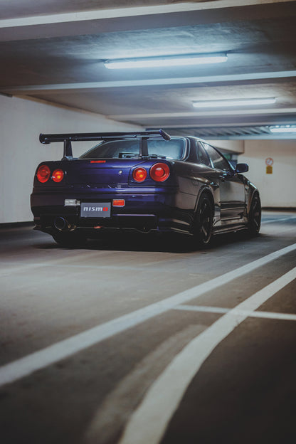 Dunkelvioletter Nissan Skyline R34 in tiefer Heck-Dreiviertelansicht in einer Beton-Parkgarage. Sichtbar sind vier rote Rundlichter, ein großer Heckflügel, dunkle Räder, ein großes Auspuffendrohr, weiße Fahrbahnlinien und kühle Deckenleuchten.