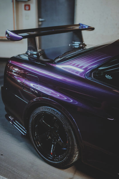 Nahaufnahme der hinteren Seitenpartie eines violetten Nissan Skyline R34 in einem Innenraum; sichtbar sind schwarzes Hinterrad, Radlauf, Heckscheibe und großer Heckflügel mit hellen Reflexionen.