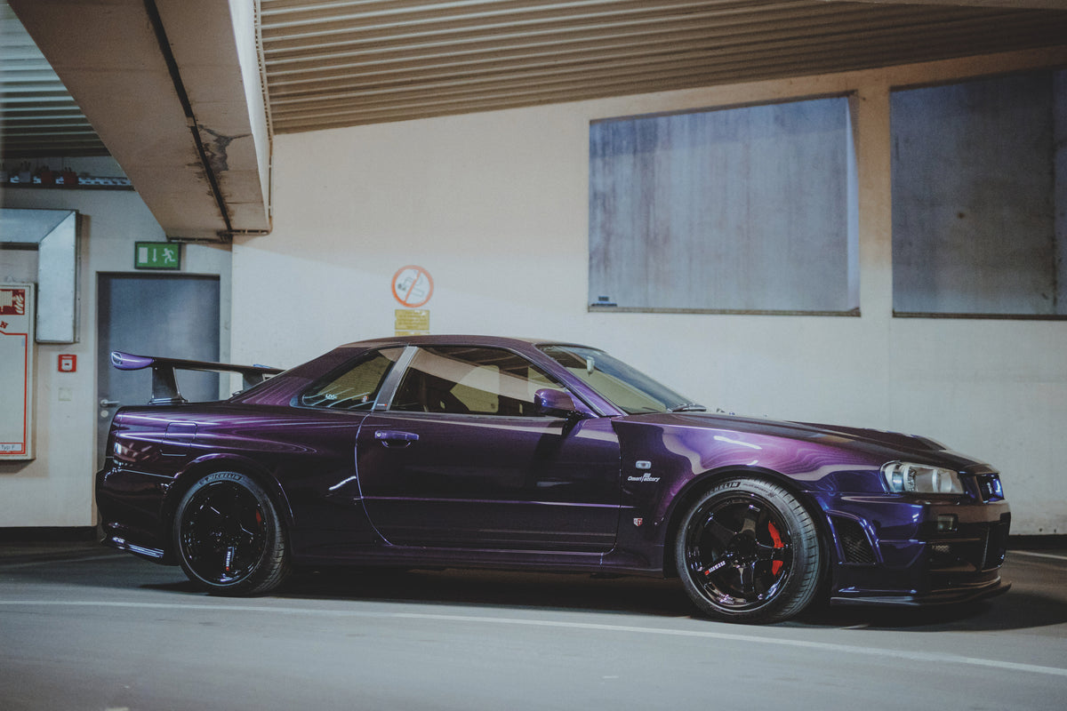Violetter Nissan Skyline R34 seitlich mit leicht sichtbarer Front vor einer hellen Garagenwand.