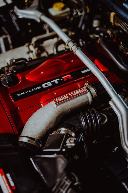 Nahaufnahme eines Motorraums mit roter Abdeckung und sichtbarem SKYLINE GT-R-Schriftzug, hellem Rohr mit TWIN TURBO-Kennzeichnung, schwarzer Verrohrung und einer silbernen, diagonal verlaufenden Strebe; dunkler Hintergrund mit warmen Lichtreflexen.