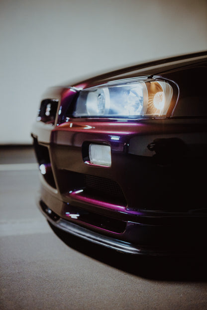 Nahaufnahme der Front eines dunklen, bordeauxviolett wirkenden Nissan Skyline V-Spec aus niedriger, schräger Perspektive. Ein rechteckiger Scheinwerfer mit kühlen und warmen Lichtanteilen dominiert die rechte Bildhälfte. Darunter sind Frontschürze, Leuchte und Lufteinlass zu sehen; eine hellgraue Wand und ein grauer Boden bilden den unscharfen Hintergrund.