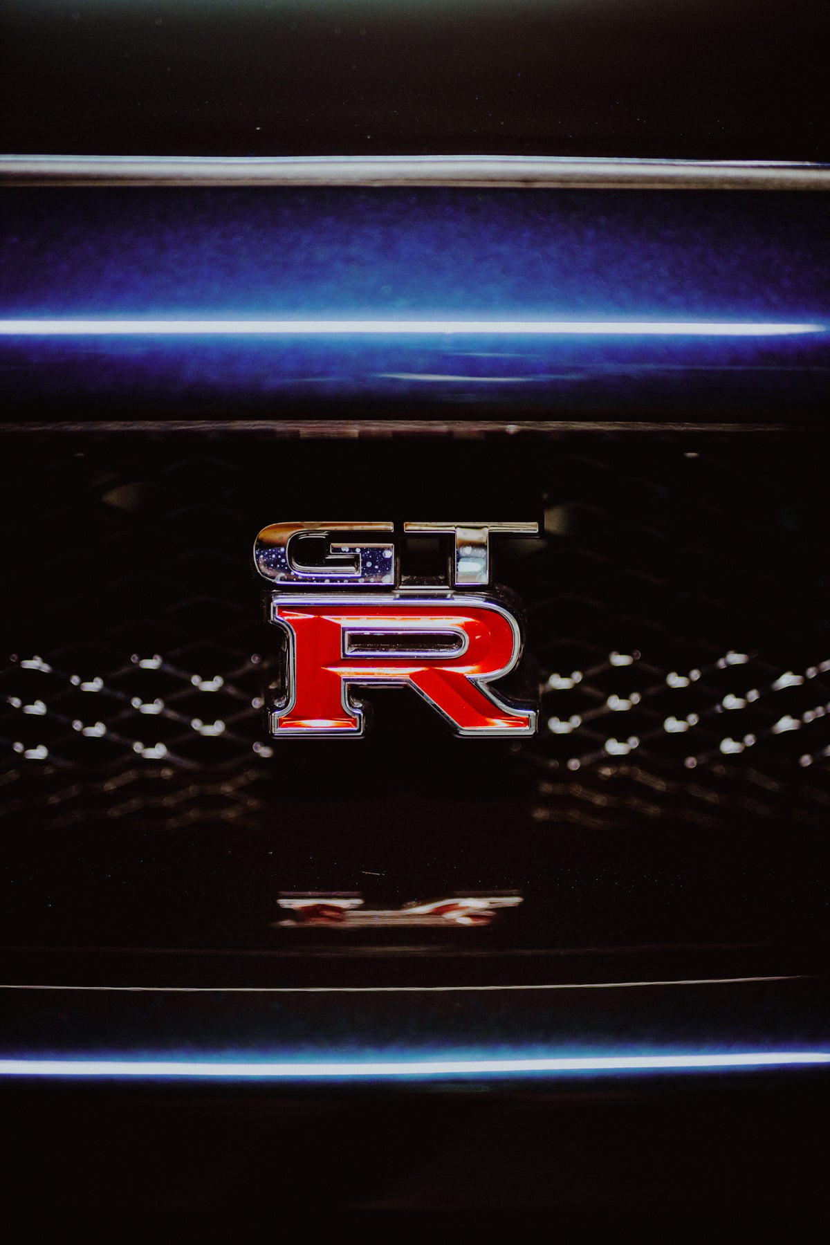 Nahaufnahme des rot-silbernen GT-R-Emblems im schwarzen Kühlergrill unter einer blauvioletten Lackkante.