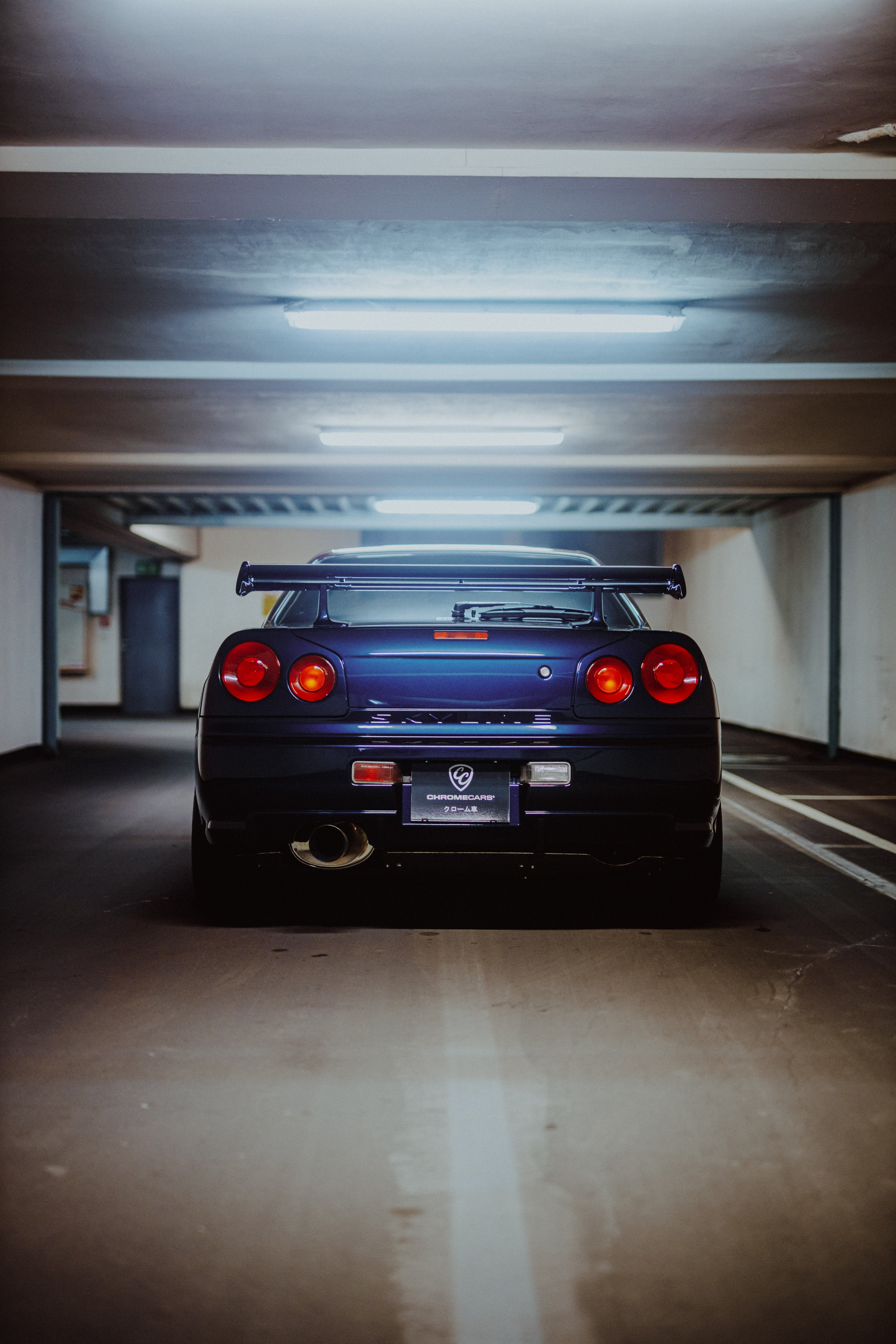 Gerade Heckansicht eines dunklen Nissan Skyline R34 in einer Tiefgarage unter mehreren Deckenleuchten.