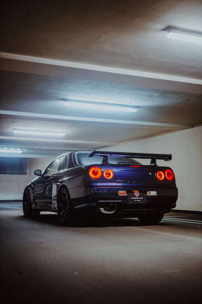 Dunkler Nissan Skyline R34 in niedriger Heck-Dreiviertelansicht mit vier rot leuchtenden Rundrückleuchten, großem Heckflügel und sichtbarem Auspuff in einer Beton-Parkgarage unter kühlen Deckenleuchten.