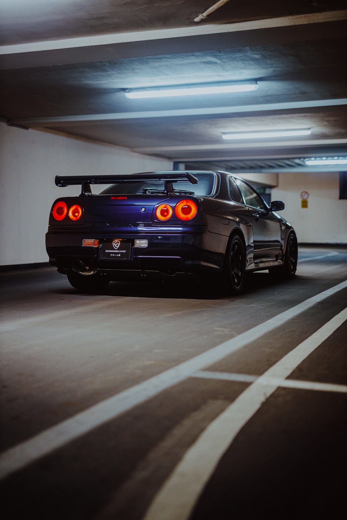 Dunkler Nissan Skyline R34 schräg von hinten mit leuchtenden runden Rückleuchten und weißen Bodenmarkierungen.