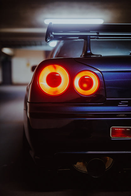 Nahaufnahme der Heckpartie eines dunklen Nissan Skyline R34 V-Spec mit zwei sichtbaren, rot-orange leuchtenden Rundrückleuchten, großem Heckflügel und rundem Auspuffendrohr vor unscharfem, kühl beleuchtetem Hintergrund.
