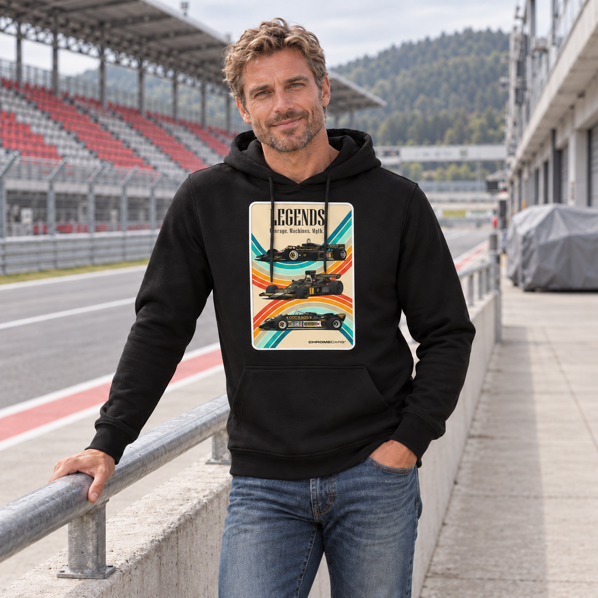 Mann im schwarzen Hoodie am Geländer einer Rennstreckentribüne. Vorn zeigt der Hoodie eine helle LEGENDS-Grafik mit drei Rennwagen und geschwungenen Farbbändern.