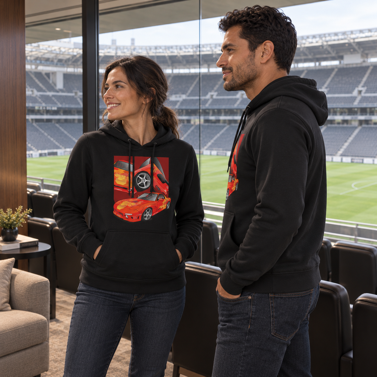 Paar in schwarzen Hoodies steht in einer Lounge mit Stadionblick. Die Frau zeigt das orangegelbe Frontmotiv, der seitlich stehende Mann eine teilweise sichtbare Seiten- und Rückansicht.