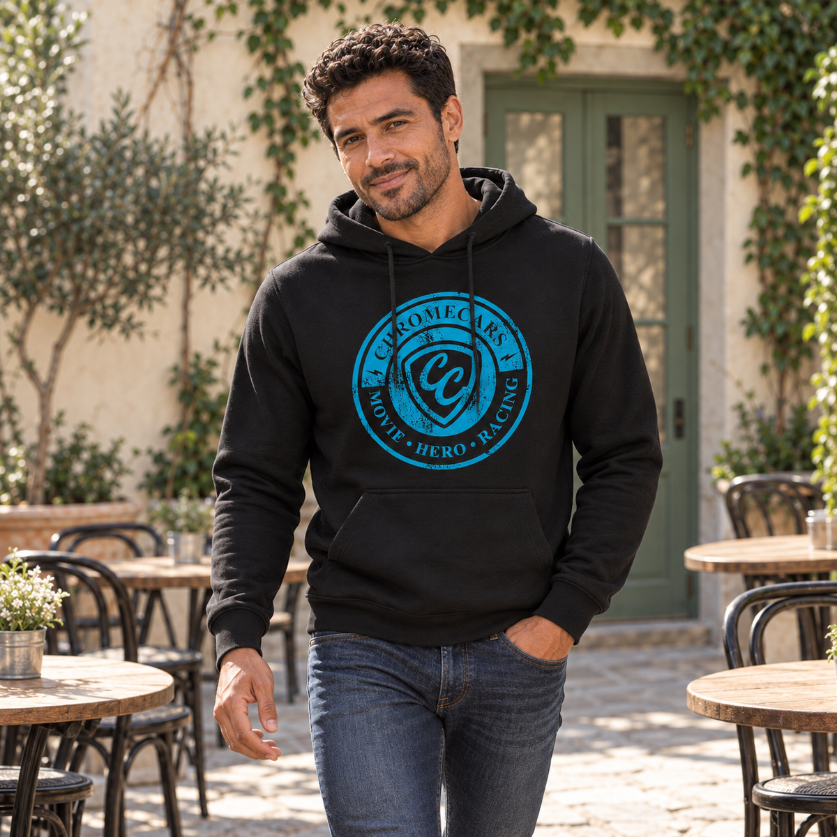 Ein Mann geht im schwarzen Hoodie mit blauem CC-Stempelmotiv zwischen den Tischen eines Café-Innenhofs. Eine Hand steckt in der Jeanstasche, der andere Arm hängt seitlich.