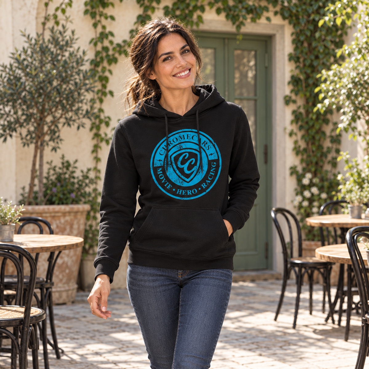 Eine Frau geht im schwarzen Hoodie mit blauem Stempelmotiv auf der Brust durch einen Café-Innenhof. Eine Hand steckt in der Kängurutasche; hinter ihr stehen Tische vor einer begrünten Wand.