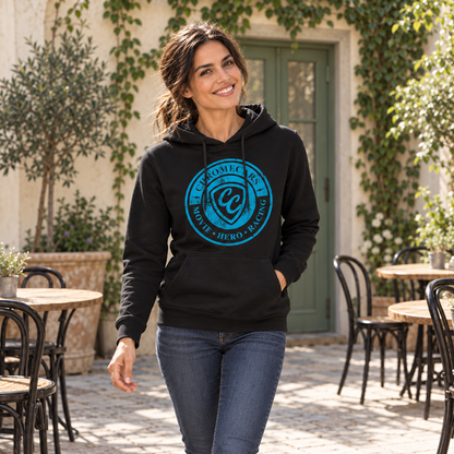 Eine Frau geht im schwarzen Hoodie mit blauem Stempelmotiv auf der Brust durch einen Café-Innenhof. Eine Hand steckt in der Kängurutasche; hinter ihr stehen Tische vor einer begrünten Wand.