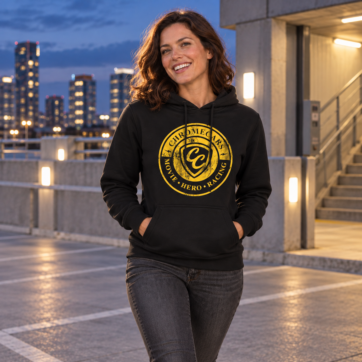 Eine Frau steht mit beiden Händen in der Kängurutasche auf einem Parkdeck neben einem beleuchteten Treppenaufgang. Ihr schwarzer Hoodie zeigt vorn das gelbe ChromeCars-Stempelmotiv.