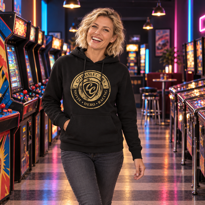 Eine Frau im schwarzen Hoodie mit goldfarbenem CC-Stempelmotiv steht mit leicht geneigtem Kopf zwischen Spielautomaten und Flippern einer Retro-Spielhalle; eine Hand steckt in der Kängurutasche.