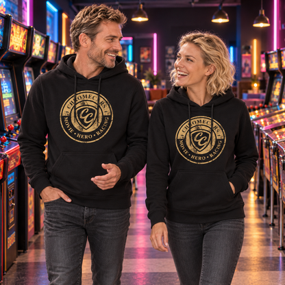 Ein Mann und eine Frau gehen in schwarzen Hoodies mit goldfarbenen CC-Stempelmotiven nebeneinander durch eine Retro-Spielhalle und sehen einander an; der Mann hält eine Hand gestikulierend vor dem Körper.