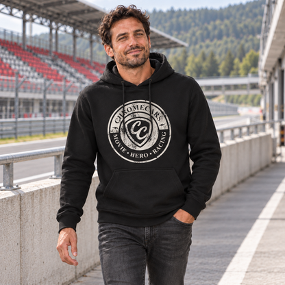 Ein Mann geht im schwarzen Hoodie mit weißer runder ChromeCars-Grafik an der Boxenmauer entlang, eine Hand in der Hosentasche; gegenüber liegt eine überdachte Rennstreckentribüne.