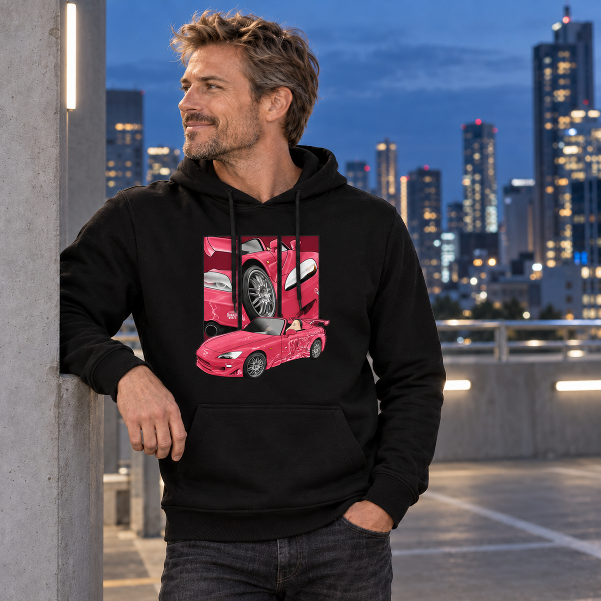 Mann im schwarzen Hoodie mit pinkem Fahrzeugmotiv lehnt an einer Betonsäule und blickt zur Seite; beleuchtete Hochhäuser im Hintergrund.