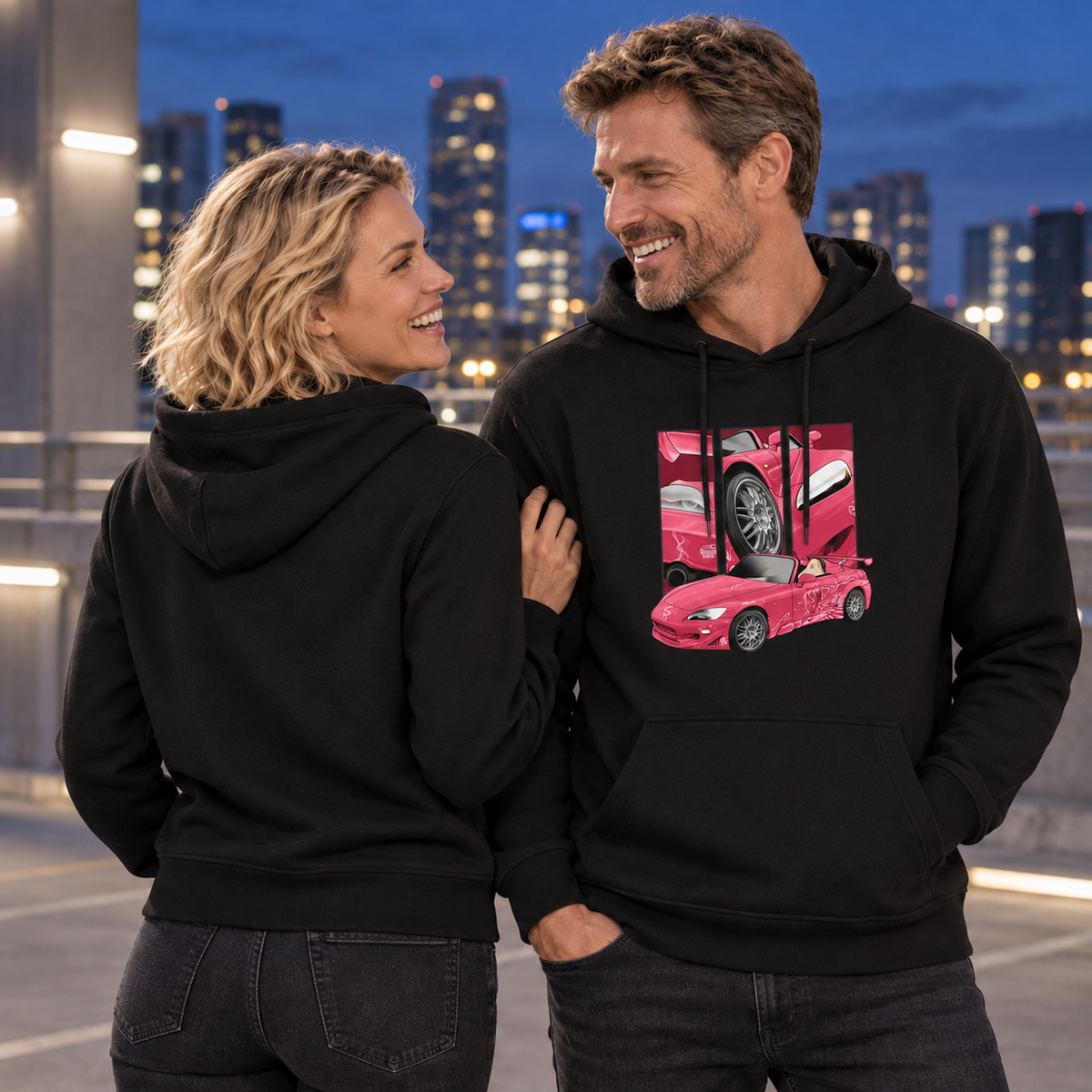 Paar in schwarzen Hoodies schaut sich lächelnd an; Frau mit unbedrucktem Rücken zur Kamera hält den Arm des Mannes, dessen Frontmotiv sichtbar ist.