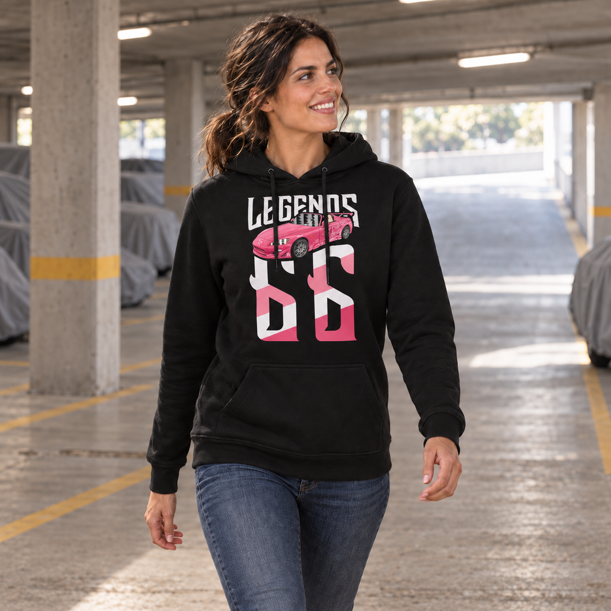 Frau im schwarzen Legends-Hoodie mit Suki’s S2000 auf der Vorderseite steht neben einer Betonsäule in der Tiefgarage und blickt zur Seite.