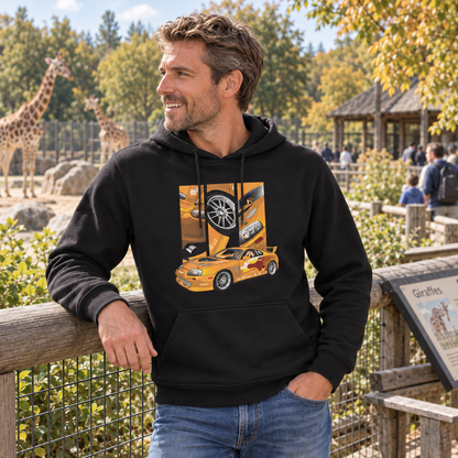Mann im schwarzen Hoodie am Holzgeländer eines Zoogeheges. Das orangefarbene Coupé mit drei Detailfeldern ist vorn sichtbar; im Hintergrund stehen Giraffen.